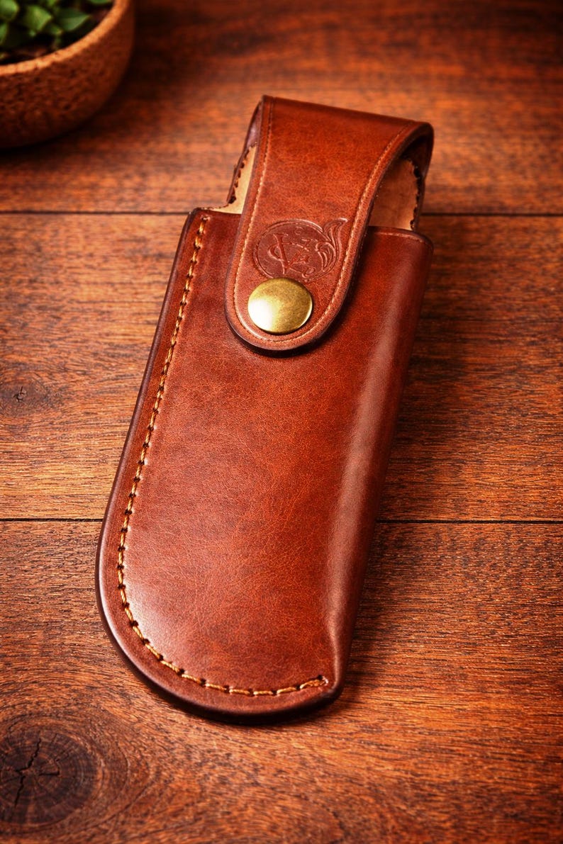 May include: A brown leather knife sheath with a flap closure and a brass button. The sheath is stitched with a contrasting thread and features a decorative emblem on the flap. The leather has a rich, warm tone.