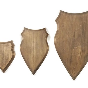 Peut inclure: Trois plaques en bois en forme de bouclier, disposées par taille croissante de gauche à droite. Chaque plaque présente une finition en bois brun clair avec un grain visible, un bord biseauté et une base pointue pour fixation murale.