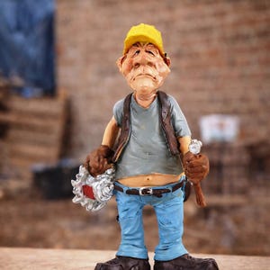 Op de afbeelding: Een beeldje van een bouwvakker met een gele helm, een blauwe spijkerbroek en een bruin vest. Hij houdt een zaag en een hamer vast. Het beeldje heeft een serieuze uitdrukking.