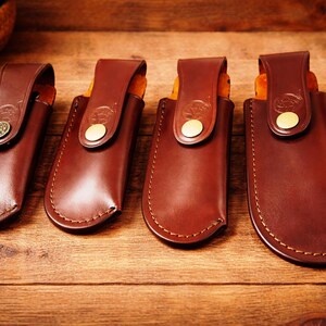 May include: Four brown leather knife sheaths with decorative stitching and brass-colored button closures. The sheaths are arranged on a wooden surface, showcasing their craftsmanship and design. The leather has a rich, warm tone.