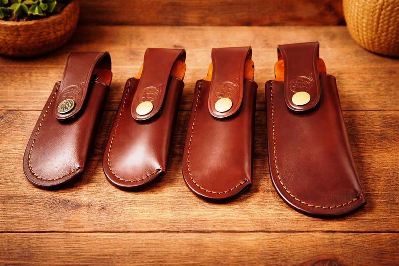 May include: Four brown leather knife sheaths with decorative stitching and brass-colored button closures. The sheaths are arranged on a wooden surface, showcasing their craftsmanship and design. The leather has a rich, warm tone.