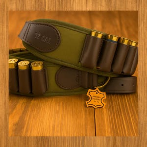 Op de afbeelding: Een olijfgroene patronengordel met bruine lederen details. De riem heeft bruine leren lussen met goudkleurige hagelpatronen. De tekst "12 CAL" is in reliëf aangebracht op een leren patch. Een leren riem en een label zijn ook zichtbaar.