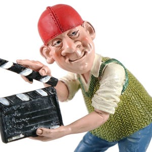 Może przedstawiać: Figurka uśmiechniętego mężczyzny w czerwonej czapce, zielonej kamizelce i niebieskich dżinsach, trzymającego klaps filmowy. Klaps jest czarny z białymi paskami i tekstem. Figurka ma szczegółowy, kreskówkowy styl.