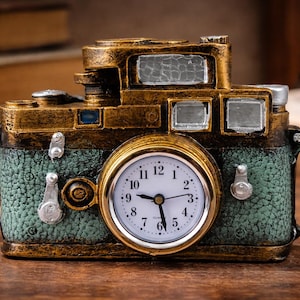 Puede incluir: Un reloj con forma de cámara de estilo vintage, con acabado dorado y verde azulado. La esfera del reloj es blanca con números y manecillas negras. La cámara tiene una superficie texturizada y está sobre madera.