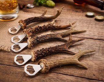 Op de afbeelding: Vijf flesopeners in de vorm van geweien met zilveren metalen openerkoppen zijn gerangschikt op een houten oppervlak. De geweien zijn bruin en beige, met een natuurlijk, rustiek uiterlijk. Een glas bier en kroonkurken staan op de achtergrond.