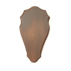 Puede incluir: Una placa de madera marrón con un borde festoneado. La placa tiene forma de escudo y tiene una superficie lisa y pulida.