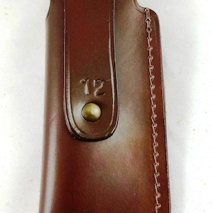 May include: A brown leather sheath with a flap secured by a brass-colored stud. The sheath has a stitched edge and the number "12" is embossed on the front. The leather has a glossy finish.