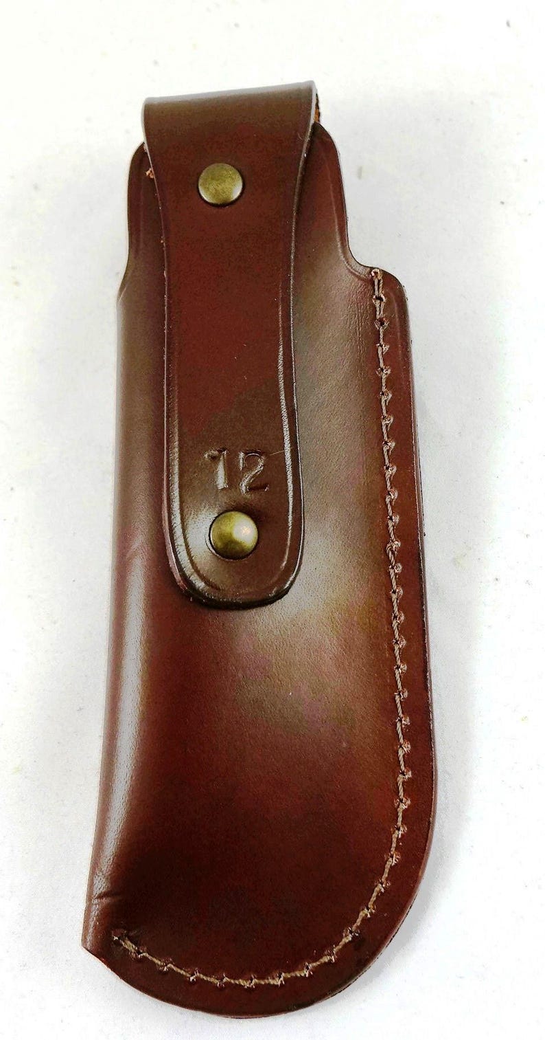 May include: A brown leather sheath with a flap secured by a brass-colored stud. The sheath has a stitched edge and the number "12" is embossed on the front. The leather has a glossy finish.