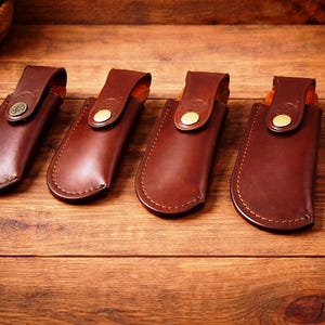May include: Four brown leather sheaths with stitched edges and brass button closures are displayed on a wooden surface. Each sheath has a flap with a button. The sheaths are designed to hold a knife or similar tool.