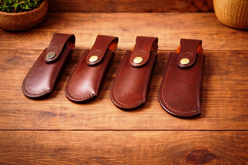 May include: Four brown leather sheaths with stitched edges and brass button closures are displayed on a wooden surface. Each sheath has a flap with a button. The sheaths are designed to hold a knife or similar tool.