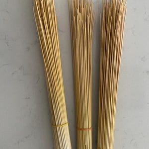 May include: Three bundles of thin, light brown wooden sticks, each held together with a rubber band. The bundles are arranged vertically against a light grey surface, creating a natural, textured appearance.