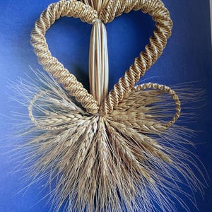 May include: A handcrafted wheat heart decoration with a bow of wheat stalks. The heart is made of woven straw and is centered on a blue background. The wheat has a natural, golden color.