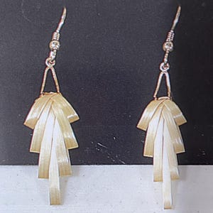 May include: A pair of drop earrings featuring a fan-shaped design made of layered, light-coloured, woven strips. Each earring hangs from a silver-toned metal hook and a triangular wire frame, creating a tiered, fringe-like appearance.