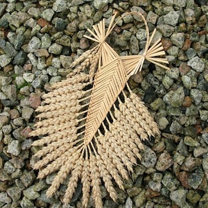 Peut inclure: Une gerbe de blé tissée avec un motif décoratif, réalisée à partir de matériaux naturels. La gerbe est présentée sur un lit de petites pierres grises.