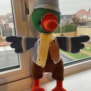 May include: A handcrafted crochet bird doll with a green head, orange beak, and a grey hat. It wears a yellow scarf, a striped sweater, and brown trousers. The bird has blue and grey wings and orange feet. A whimsical, handmade item.