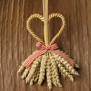 May include: A heart-shaped wheat wreath with a red and white gingham ribbon. The wreath is made of dried wheat stalks and is hanging on a wooden background.