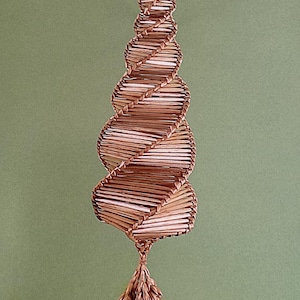 Puede incluir: Un adorno de color marrón, tejido, en forma de espiral con una borla larga de color marrón que cuelga de la parte inferior. El adorno está hecho de paja y recuerda a un árbol de Navidad.