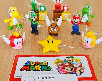 May include: A collection of Super Mario character figurines, including Mario, Luigi, Yoshi, and Bowser. The figurines are yellow, green, red, and brown. The figurines are on a wooden surface with a Super Mario themed card.