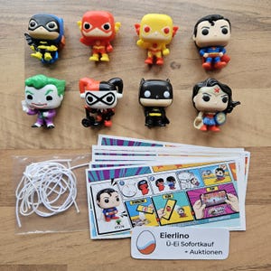 May include: A collection of eight small plastic figurines of DC Comics characters, including Batman, Superman, Wonder Woman, The Flash, Harley Quinn, The Joker, and Batgirl. The figurines are arranged in two rows of four on a wooden surface. There are also several small cards with instructions on how to play a game with the figurines.