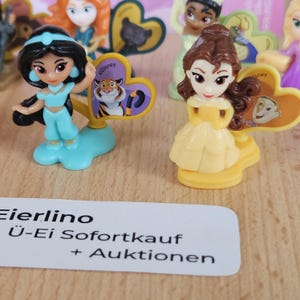 May include: Two small plastic figurines of Disney princesses, Jasmine and Belle, with their respective cartoon characters on the base. Jasmine is wearing a blue outfit and Belle is wearing a yellow dress.
