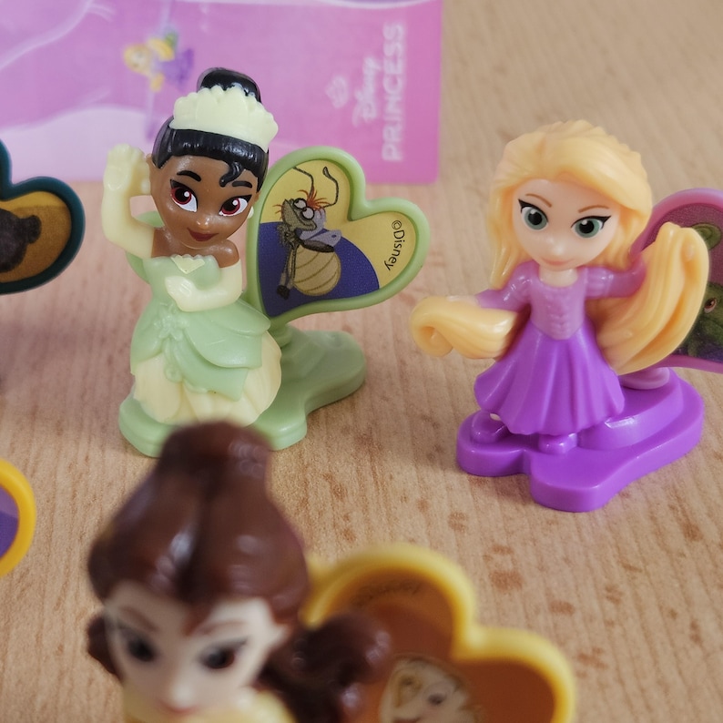 May include: Three Disney Princess figurines, Tiana, Rapunzel, and Belle, with their signature colors and accessories. Each figurine has a heart-shaped base with a Disney logo.