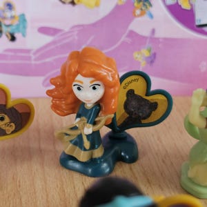 May include: A red-haired Disney princess figurine holding a bow and arrow. She is wearing a green dress with gold accents. The figurine is standing on a green base with a heart-shaped yellow sign that says "Disney" and has a black bear on it.