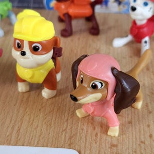May include: Two plastic toy dogs from the Paw Patrol cartoon. The dog on the left is brown and white with a yellow construction hat and vest. The dog on the right is brown with a pink helmet and pink jumpsuit.