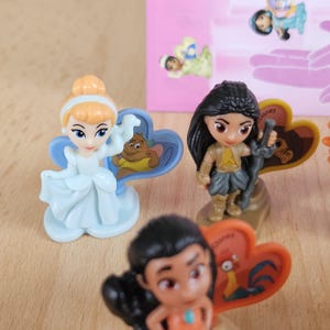 May include: A set of Disney character figurines, including Cinderella, Pocahontas, and Mulan. Each figurine is posed in a unique way and comes with a heart-shaped base featuring a Disney character. The figurines are made of plastic and are approximately 2 inches tall.
