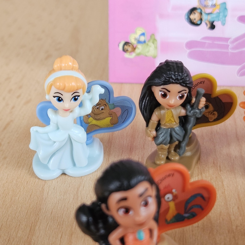 May include: A set of Disney character figurines, including Cinderella, Pocahontas, and Mulan. Each figurine is posed in a unique way and comes with a heart-shaped base featuring a Disney character. The figurines are made of plastic and are approximately 2 inches tall.
