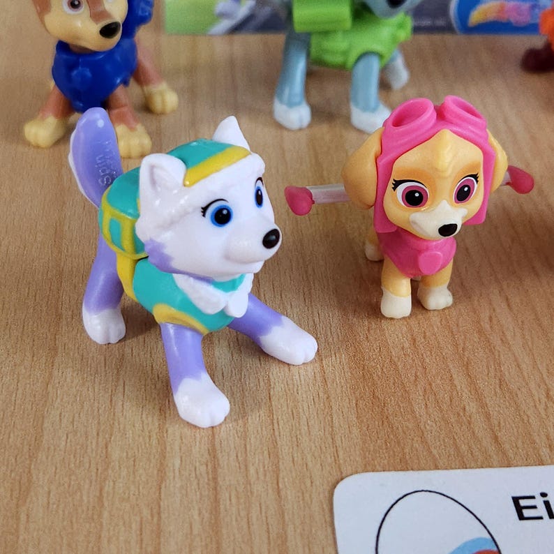 May include: Two small plastic toy dogs from the Paw Patrol cartoon. The dog on the left is white with purple legs and a teal and yellow backpack. The dog on the right is yellow with pink goggles and a pink backpack.