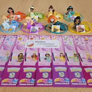 May include: Eight colorful plastic rings with Disney Princess figurines attached. Each ring has a heart-shaped charm with a princess image. The rings are displayed on a wooden surface with pink and white cards that say "Download and Play" and "Kinder.com/app".