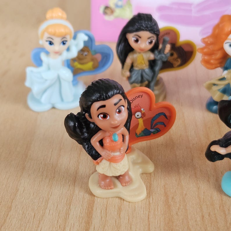 May include: A small plastic figurine of Moana from Disney's Moana. She is wearing a brown and orange dress and a blue necklace. She is standing on a brown base with a heart-shaped red and orange sign that says "Disney" and has a rooster on it.