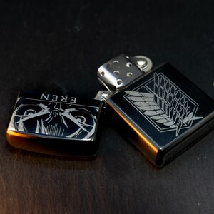 Puede incluir: Dos encendedores de metal negro con grabados plateados. Un encendedor presenta la palabra "EREN" y un dise&ntilde;o de personaje. El otro encendedor tiene un dise&ntilde;o de ala y emblema. Ambos encendedores est&aacute;n abiertos y expuestos sobre una superficie oscura.