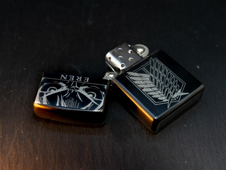 Puede incluir: Dos encendedores de metal negro con grabados plateados. Un encendedor presenta la palabra "EREN" y un dise&ntilde;o de personaje. El otro encendedor tiene un dise&ntilde;o de ala y emblema. Ambos encendedores est&aacute;n abiertos y expuestos sobre una superficie oscura.