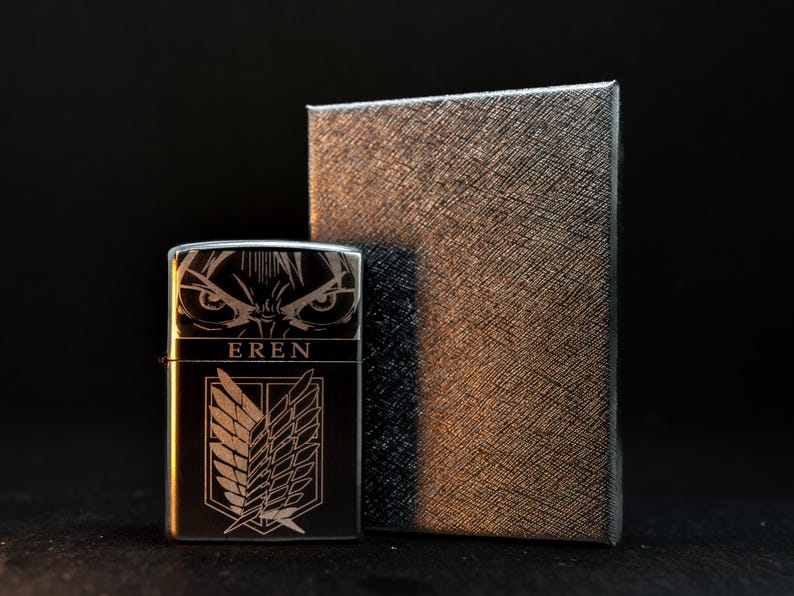 Puede incluir: Un encendedor de metal negro con el nombre "EREN" y un dise&ntilde;o gr&aacute;fico. El encendedor est&aacute; junto a una caja texturizada. El dise&ntilde;o incluye un par de ojos y un emblema de ala.