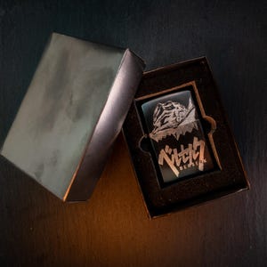 May include: A black lighter with a detailed graphic and Japanese text inside a black box. The lighter features a design with a character and the word "BERSERK". The box is open, revealing the lighter.