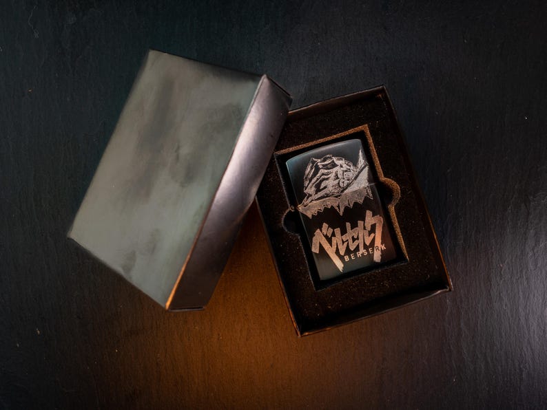 May include: A black lighter with a detailed graphic and Japanese text inside a black box. The lighter features a design with a character and the word "BERSERK". The box is open, revealing the lighter.