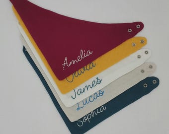 May include: Five personalized baby bibs in different colors, stacked on top of each other. The bibs are made of a soft, absorbent fabric and have a snap closure. The names Amelia, Olivia, James, Lucas, and Sophia are embroidered on the bibs in blue thread.
