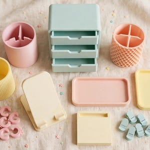 May include: A collection of pastel desk accessories including a light blue three-drawer organiser, two cylindrical pen holders in pink and yellow, a textured peach pen holder, two rectangular trays, a phone stand, and small pink and blue clips.