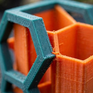 May include: A 3D printed desk organiser featuring a teal hexagonal outer frame and bright orange inner compartments. The structure displays visible layer lines from the additive manufacturing process, creating a textured surface on the geometric walls.