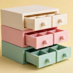 May include: A stack of three rectangular storage organisers in cream, light pink, and sage green. Each unit features two pull-out drawers with small round knobs, and each drawer is divided into six small square compartments for organised storage.