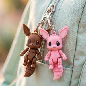May include: Two articulated keychains attached to a metal ring, featuring one brown and one pink creature with large pointed ears, round eyes, and segmented tails. The figures have movable joints at the shoulders, elbows, hips, and knees.