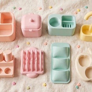 May include: A collection of eight pastel-coloured silicone organisers in peach, pink, mint green, and yellow. The set includes various shapes featuring compartments, slots, and a curved handle, designed for holding small items in a tidy arrangement.