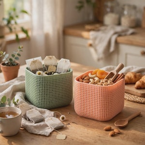 May include: Two square storage containers with a grid-patterned exterior. One container is sage green and holds several tea bags, while the other is peach-colored and contains a mix of nuts, dried apricots, and a small wooden scoop.