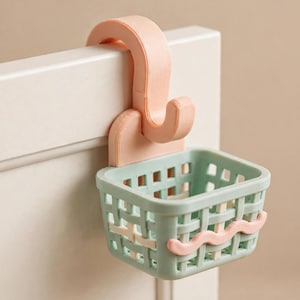 May include: A light peach-coloured over-the-door hook attached to a small, rectangular, light green basket with a grid-like woven pattern. The basket features a single wavy light peach-coloured decorative line across the front exterior surface.