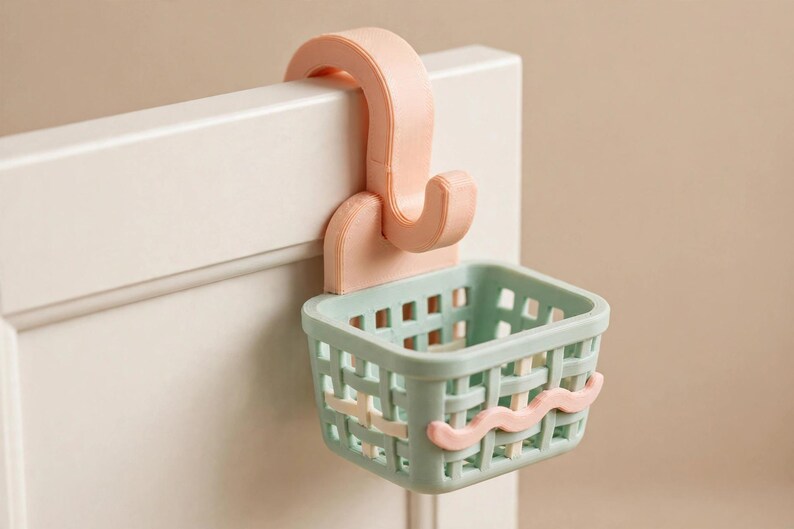 May include: A light peach-coloured over-the-door hook attached to a small, rectangular, light green basket with a grid-like woven pattern. The basket features a single wavy light peach-coloured decorative line across the front exterior surface.