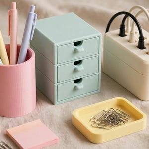 May include: A pink cylindrical pen holder containing four pastel pens, a light green three-drawer mini organiser, a cream-coloured cable management box with black and white cords, a yellow rectangular tray with metal paper clips, and sticky notes.
