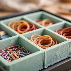 May include: A light green desk organiser with six square compartments containing various office supplies. The compartments hold small colourful rubber bands, pink, yellow, and blue square erasers, and assorted metal paper clips in pink and blue.