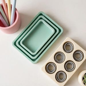 May include: A pink cylindrical pen holder containing four pastel pens, three nested rectangular sage green trays, a cream-coloured rectangular tray with six circular indentations holding grey rings, and a small cream pot with a green succulent.