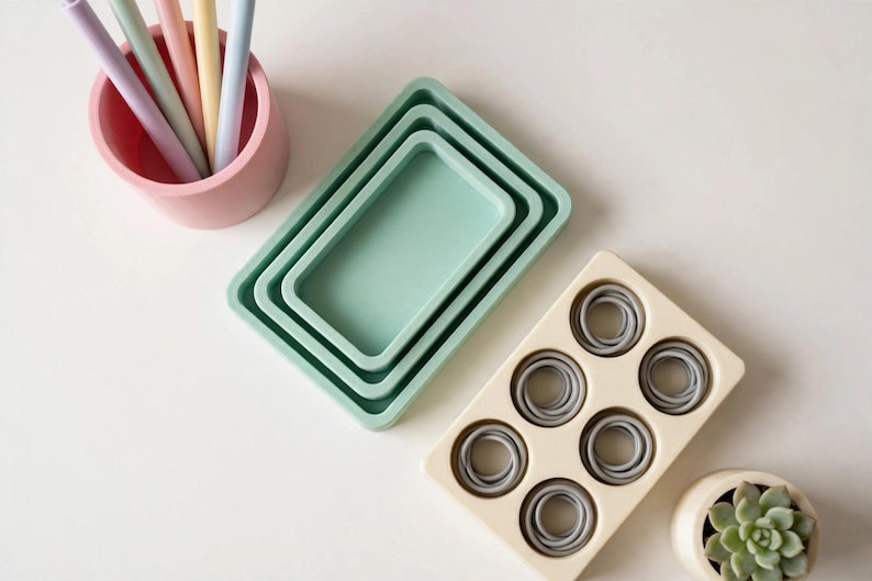 May include: A pink cylindrical pen holder containing four pastel pens, three nested rectangular sage green trays, a cream-coloured rectangular tray with six circular indentations holding grey rings, and a small cream pot with a green succulent.